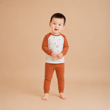 A toddler standing barefoot against a solid beige studio background, wearing a long-sleeve raglan pajama top with a white body and brown sleeves, decorated with small animal illustrations, paired with matching brown pants.