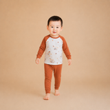A toddler standing barefoot against a solid beige studio background, wearing a long-sleeve raglan shirt with a white body and brown sleeves, plus matching brown pants.