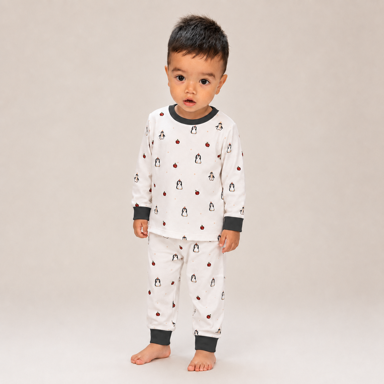 A toddler standing barefoot against a plain light beige studio background, wearing a white long-sleeve pajama top and matching pants with dark gray ribbed collar, cuffs, and hem, plus a small penguin print.