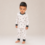 A toddler standing barefoot against a plain light beige studio background, wearing a white long-sleeve pajama top and matching pants with dark gray ribbed collar, cuffs, and hem, plus a small penguin print.