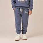A child wearing a matching slate blue sweatshirt and jogger pants set, paired with white sneakers, standing against a solid beige studio background.