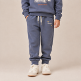 A child wearing a matching slate blue sweatshirt and jogger pants set, paired with white sneakers, standing against a solid beige studio background.