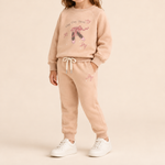 A child modeling a matching beige sweatshirt and jogger pants set with ballet-themed graphics, including a pair of pink and black pointe shoes and the words 'Tutu Tutu Ballet' on the sweatshirt.