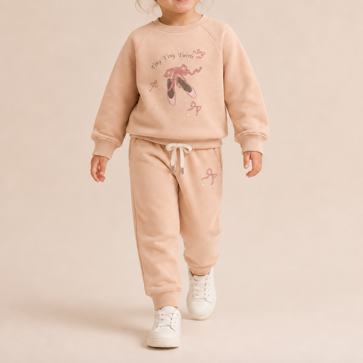 A toddler modeling a matching two-piece sweatshirt and jogger pants set in a soft peach-beige color. The sweatshirt features a graphic of a ballerina with the text 'Happy Tong Tongs' and ballet shoes, while the joggers have a drawstring waist and a small ballet shoe graphic on the thigh.