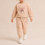 A toddler modeling a matching two-piece sweatshirt and jogger pants set in a soft peach-beige color. The sweatshirt features a graphic of a ballerina with the text 'Happy Tong Tongs' and ballet shoes, while the joggers have a drawstring waist and a small ballet shoe graphic on the thigh.