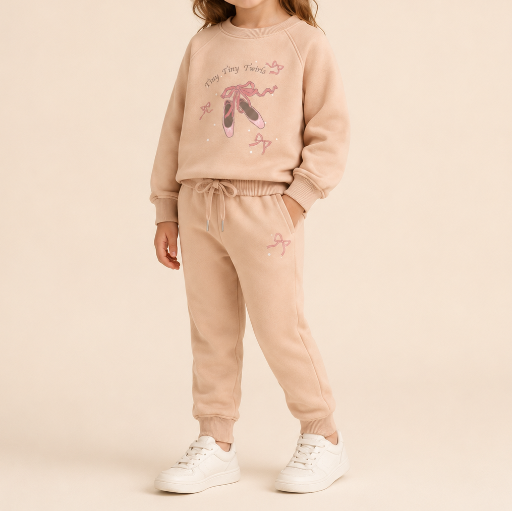 A child wearing a matching beige sweatshirt and jogger pants set with ballet-themed graphics, paired with white sneakers, against a solid beige studio background.