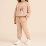 A child wearing a matching beige sweatshirt and jogger pants set with ballet-themed graphics, paired with white sneakers, against a solid beige studio background.