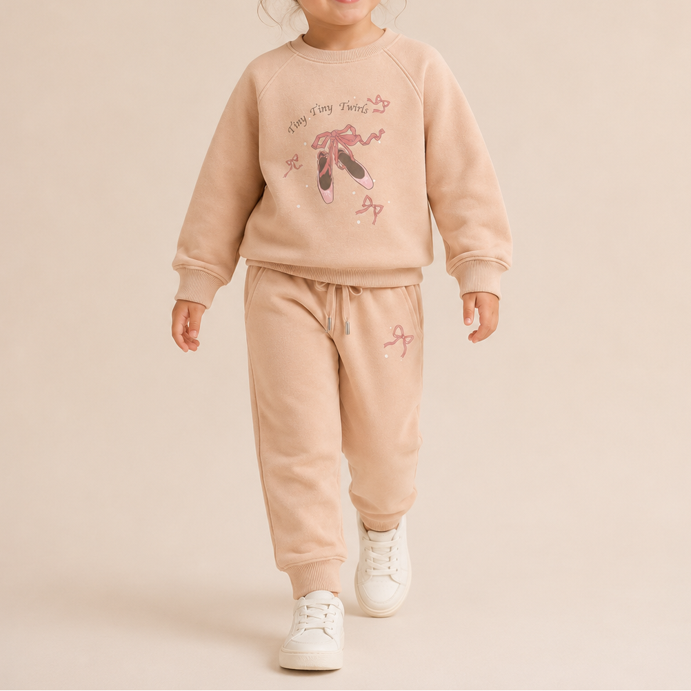 A young girl with her hair in a bun is smiling and walking forward, wearing a matching light peach-beige two-piece lounge set with a ballet theme graphic on the sweatshirt and white sneakers, set against a solid light beige studio background.