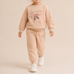 A young girl with her hair in a bun is smiling and walking forward, wearing a matching light peach-beige two-piece lounge set with a ballet theme graphic on the sweatshirt and white sneakers, set against a solid light beige studio background.