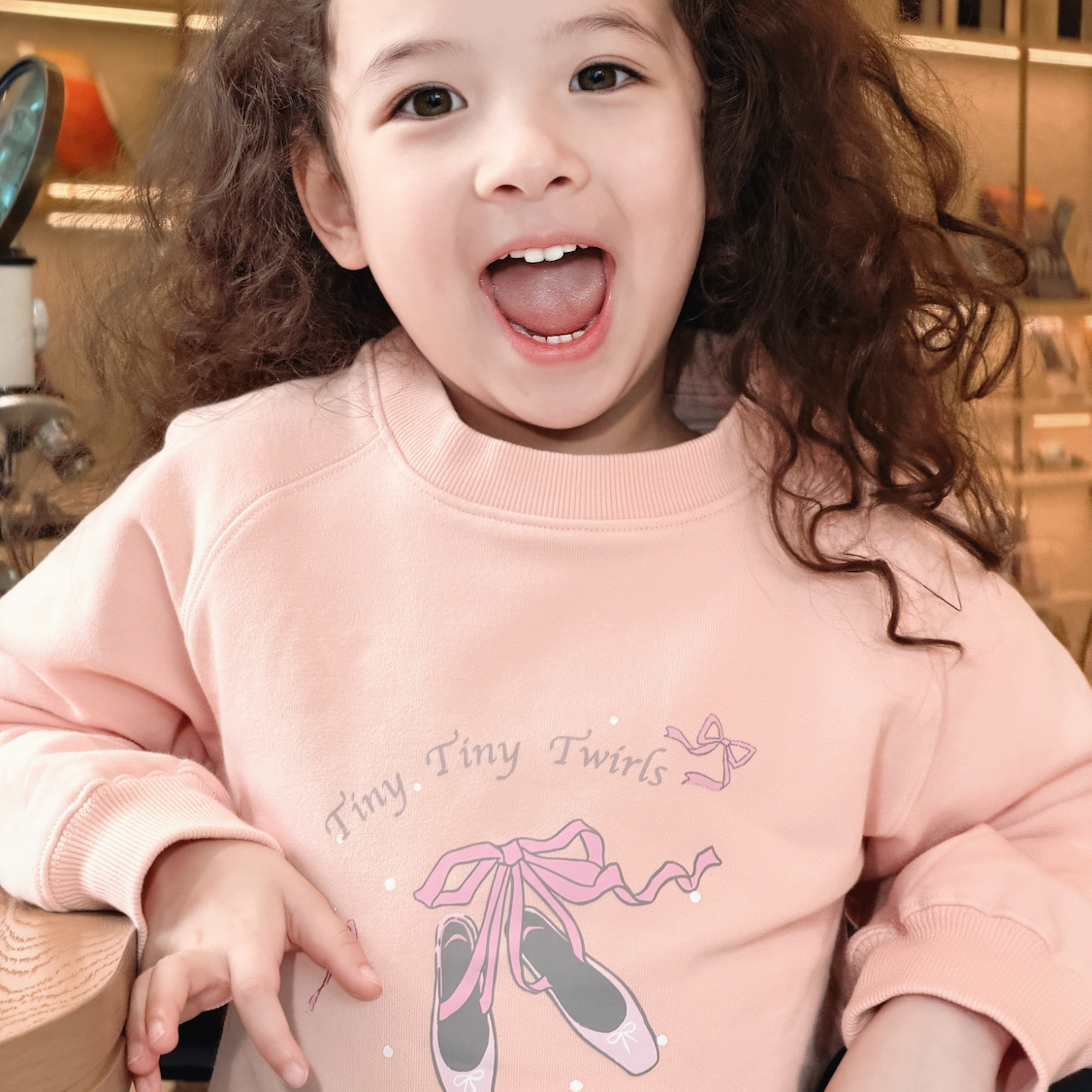 A young child with curly brown hair wearing a light pink sweatshirt with a ballet shoe graphic and the text 'Tiny, Tiny Twins' on the front, photographed in a warm indoor setting.