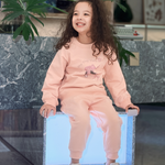 A young girl with long curly brown hair sits smiling on a glowing white block in a modern indoor setting. She is wearing a light pink two-piece sweatshirt and sweatpants set with a small graphic on the chest.