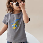 Child wearing a gray t-shirt with a graphic design and striped pants, holding sunglasses.