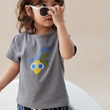 Child wearing a gray t-shirt with a graphic design and striped pants, holding sunglasses.