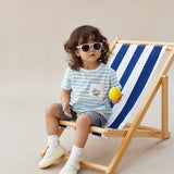 Child sitting on a striped deck chair holding a lemon, wearing sunglasses and a striped shirt.