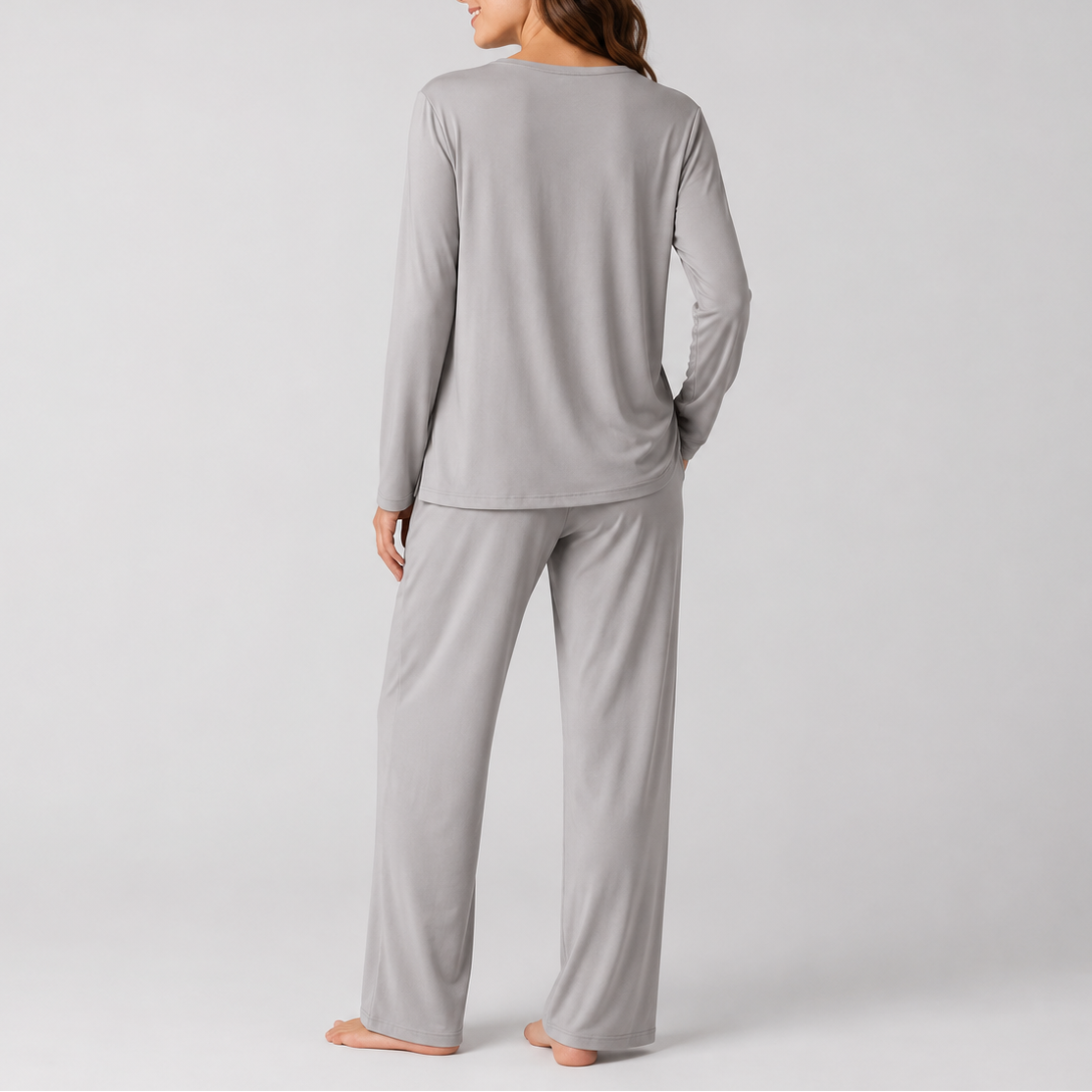 A woman shown from the back wearing a light gray long-sleeve pajama top and matching wide-leg pajama pants, standing barefoot against a plain light gray studio background.