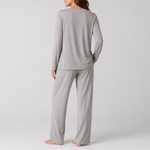 A woman shown from the back wearing a light gray long-sleeve pajama top and matching wide-leg pajama pants, standing barefoot against a plain light gray studio background.