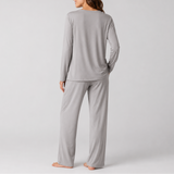 A woman shown from the back wearing a light gray long-sleeve pajama top and matching wide-leg pajama pants, standing barefoot against a plain light gray studio background.