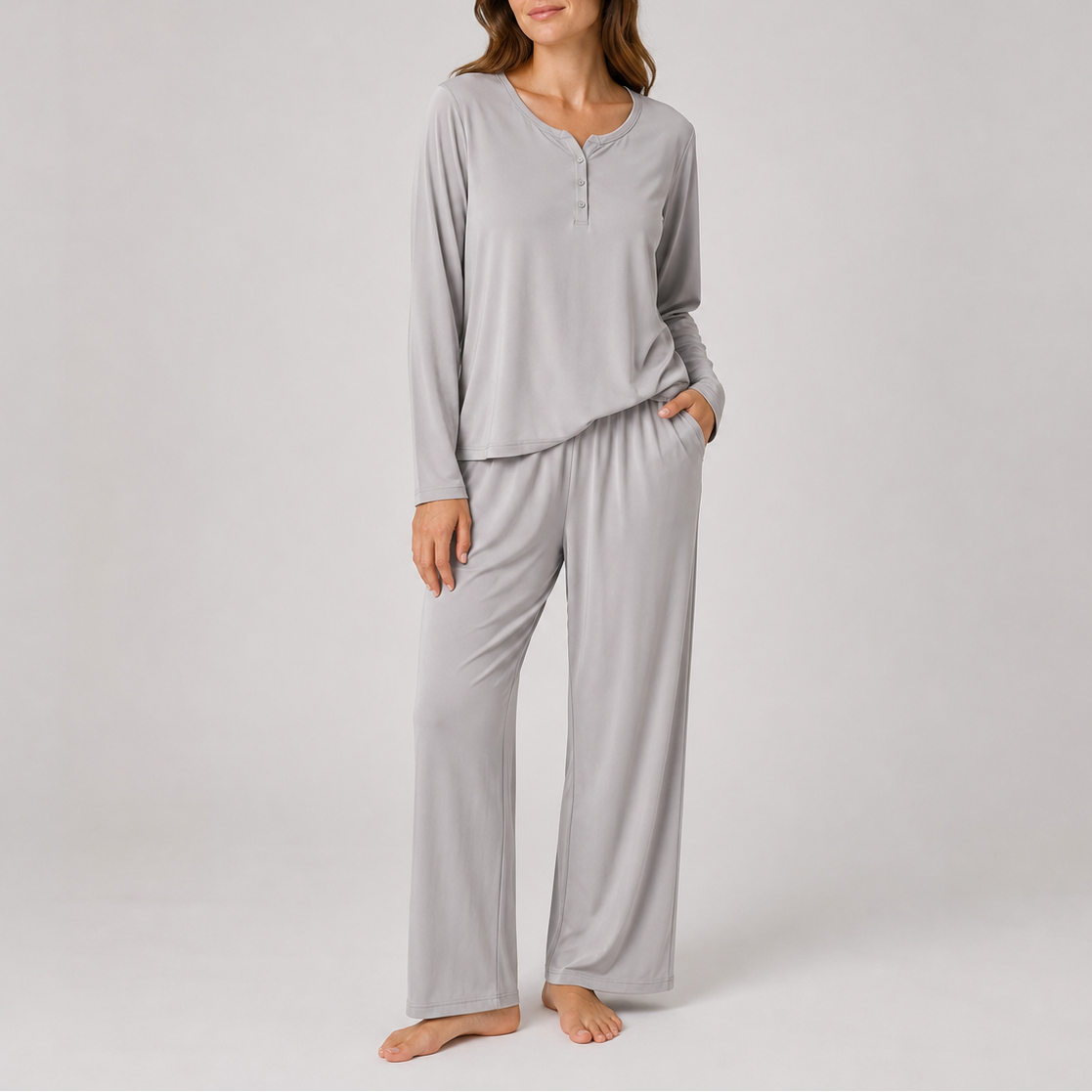 A woman modeling a light gray two-piece pajama set consisting of a long-sleeve henley top and matching wide-leg pants, photographed against a neutral light gray studio background.