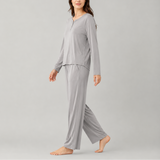 A woman modeling a light gray two-piece pajama set consisting of a long-sleeve henley top and matching wide-leg pants, photographed against a solid light gray studio background.