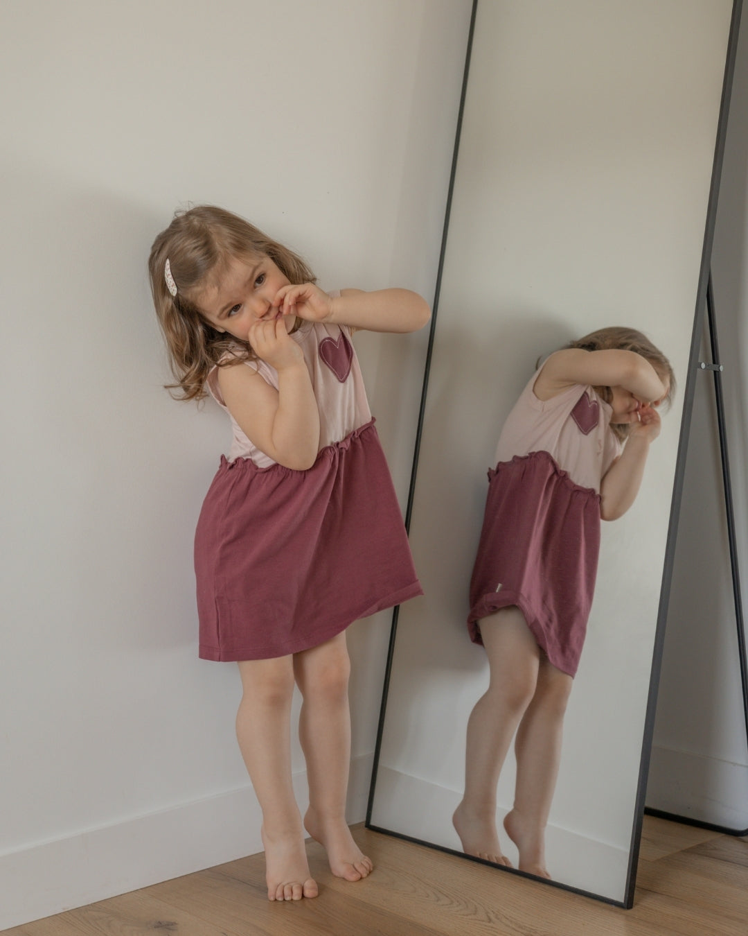 Young girl in a pink dress standing next to a mirror reflection.