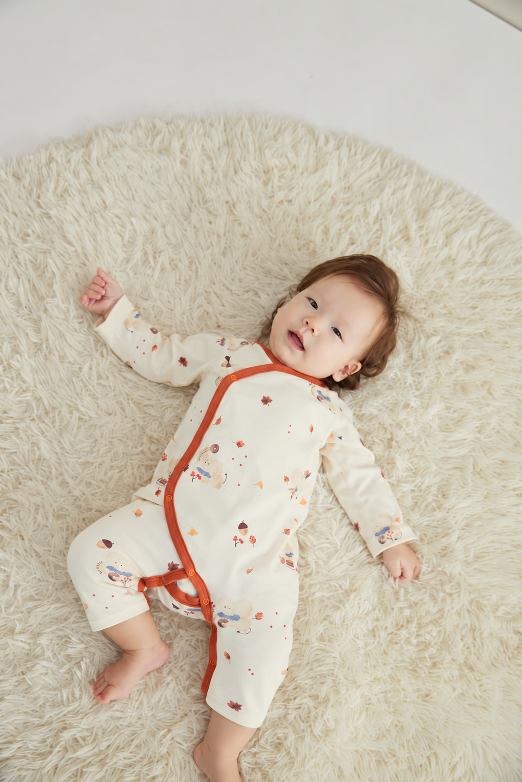 A baby lying on a fluffy cream rug, wearing a cream-colored long-sleeve footless sleeper with orange trim and small multicolor cartoon prints.
