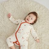 A baby lying on a fluffy cream rug, wearing a cream-colored long-sleeve footless sleeper with orange trim and small multicolor cartoon prints.