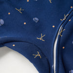 Close-up of a navy blue fabric garment with a white zipper and small gold and light blue printed motifs, including abstract shapes and a small skull-and-crossbones style design.