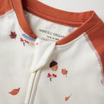 Close-up of a children's zip up bodysuit by Norsu Organic, featuring a cream body with a small autumn leaf and acorn print, and a rust-orange collar and sleeve cuffs.
