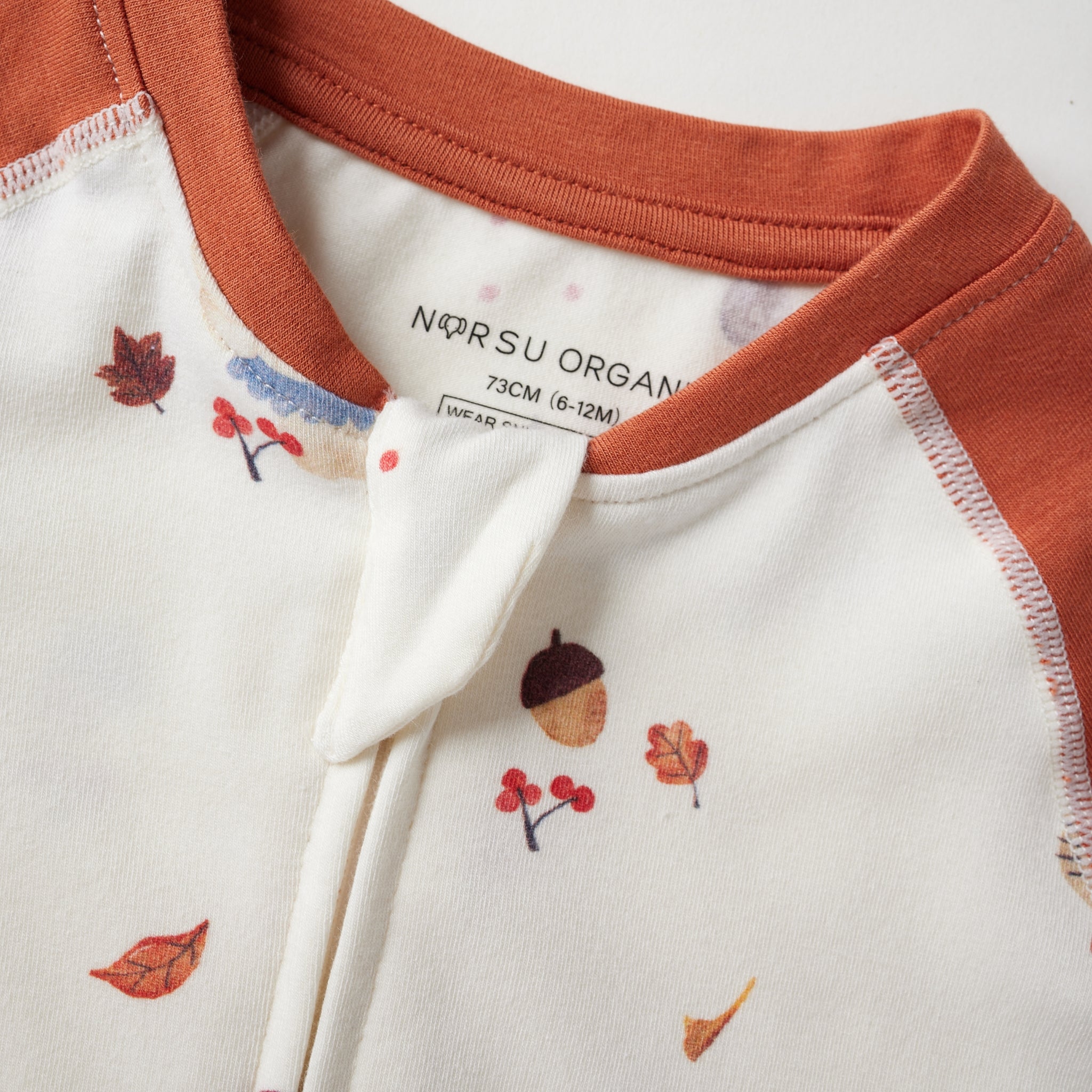 Close-up of a children's zip up bodysuit by Norsu Organic, featuring a cream body with a small autumn leaf and acorn print, and a rust-orange collar and sleeve cuffs.