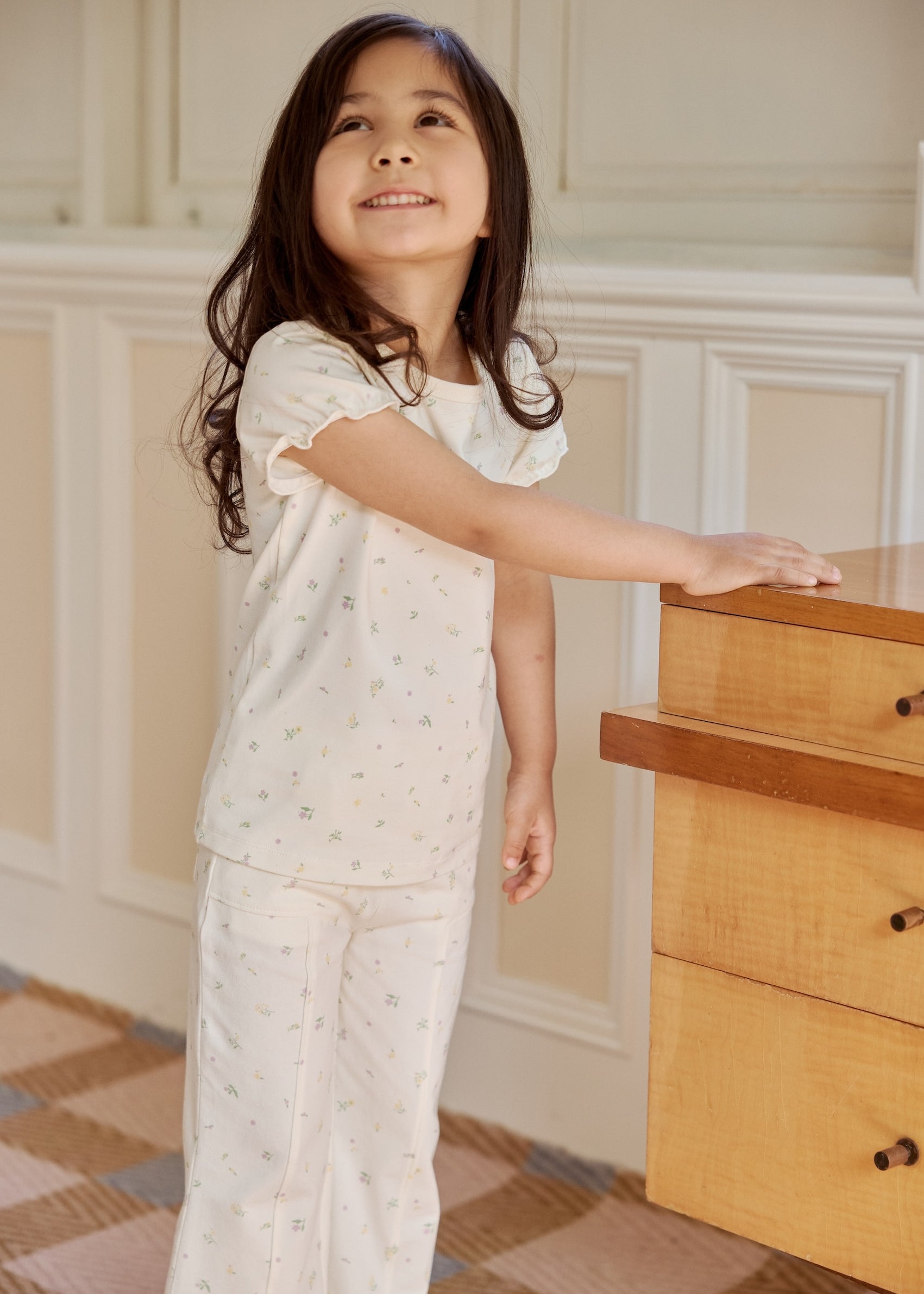 A little girl in colorful pajamas stands beside a wooden dresser, smiling and looking playful