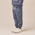 A child wearing slate blue sweatpants with white drawstrings and small embroidered details, paired with a matching sweatshirt and white sneakers, against a solid beige background.