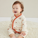 A baby sitting on a fluffy cream rug wearing a cream-colored long-sleeve romper with orange trim and small multicolor printed motifs.