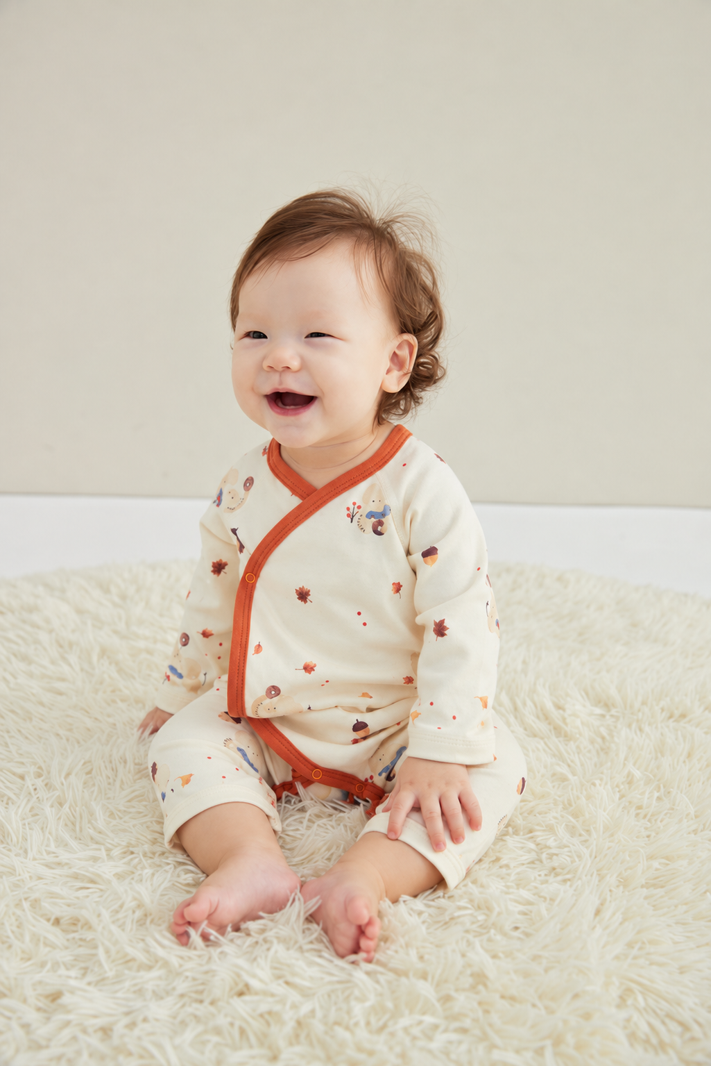 A baby sitting on a fluffy cream rug wearing a cream-colored long-sleeve romper with orange trim and small multicolor printed motifs.