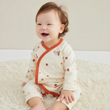 A baby sitting on a fluffy cream rug wearing a cream-colored long-sleeve romper with orange trim and small multicolor printed motifs.