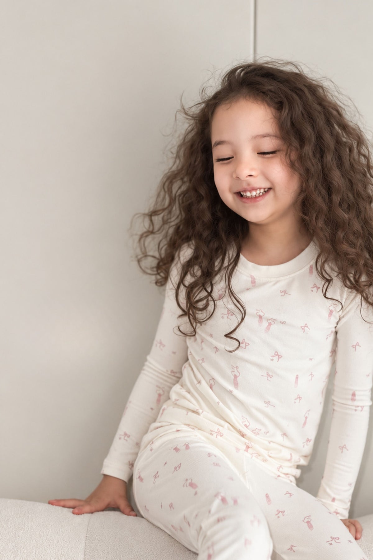A young girl with long, curly brown hair is smiling while wearing a cream-colored long-sleeve pajama top and matching pants with a small pink pattern, sitting on a light-colored cushion against a neutral wall.