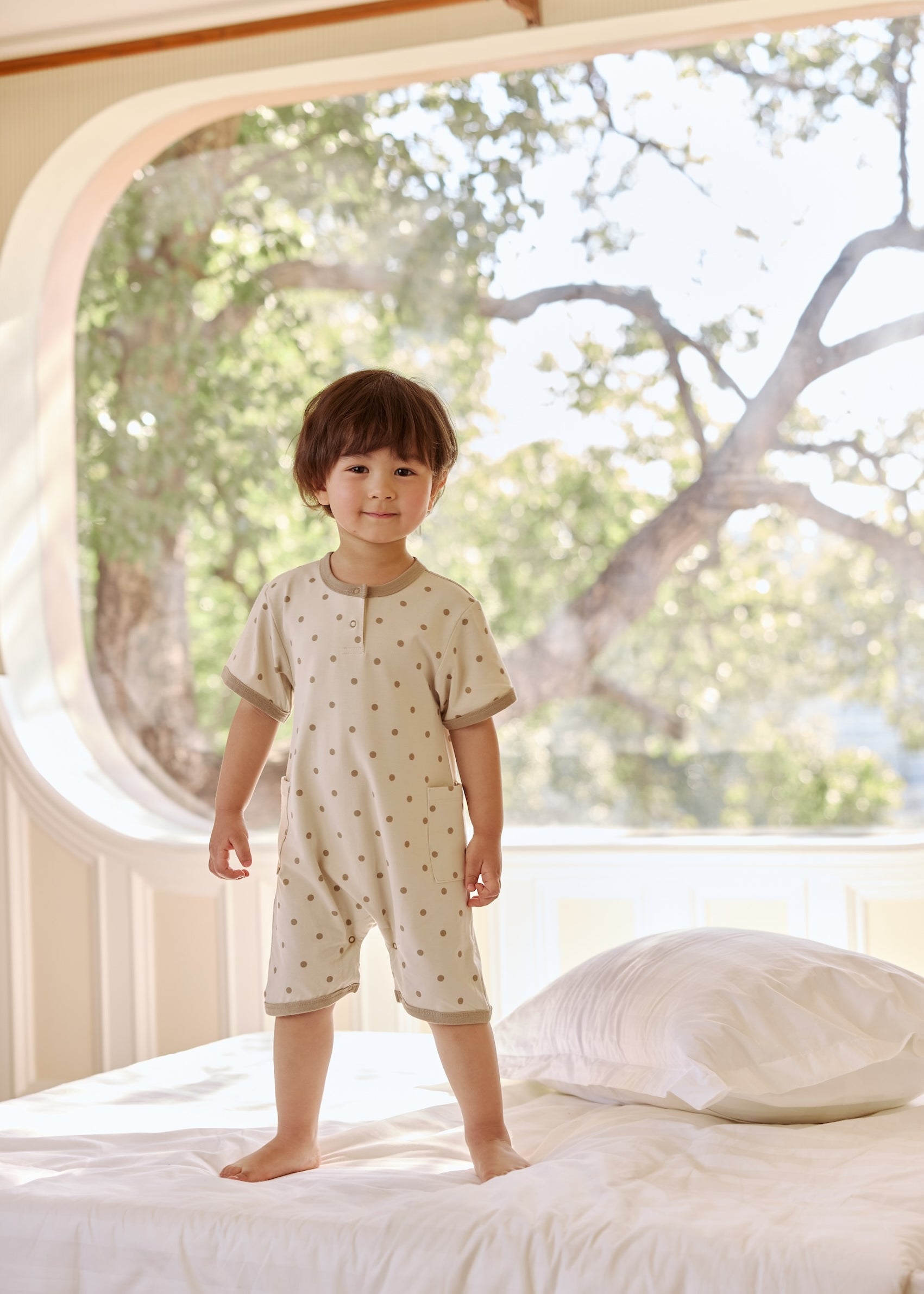 A young child standing on a bed in a bright room, wearing a polka-dot outfit, with large windows and trees outside
