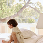 A young child sitting on a bed in a bright room, wearing a polka-dot outfit, with large windows and trees outside