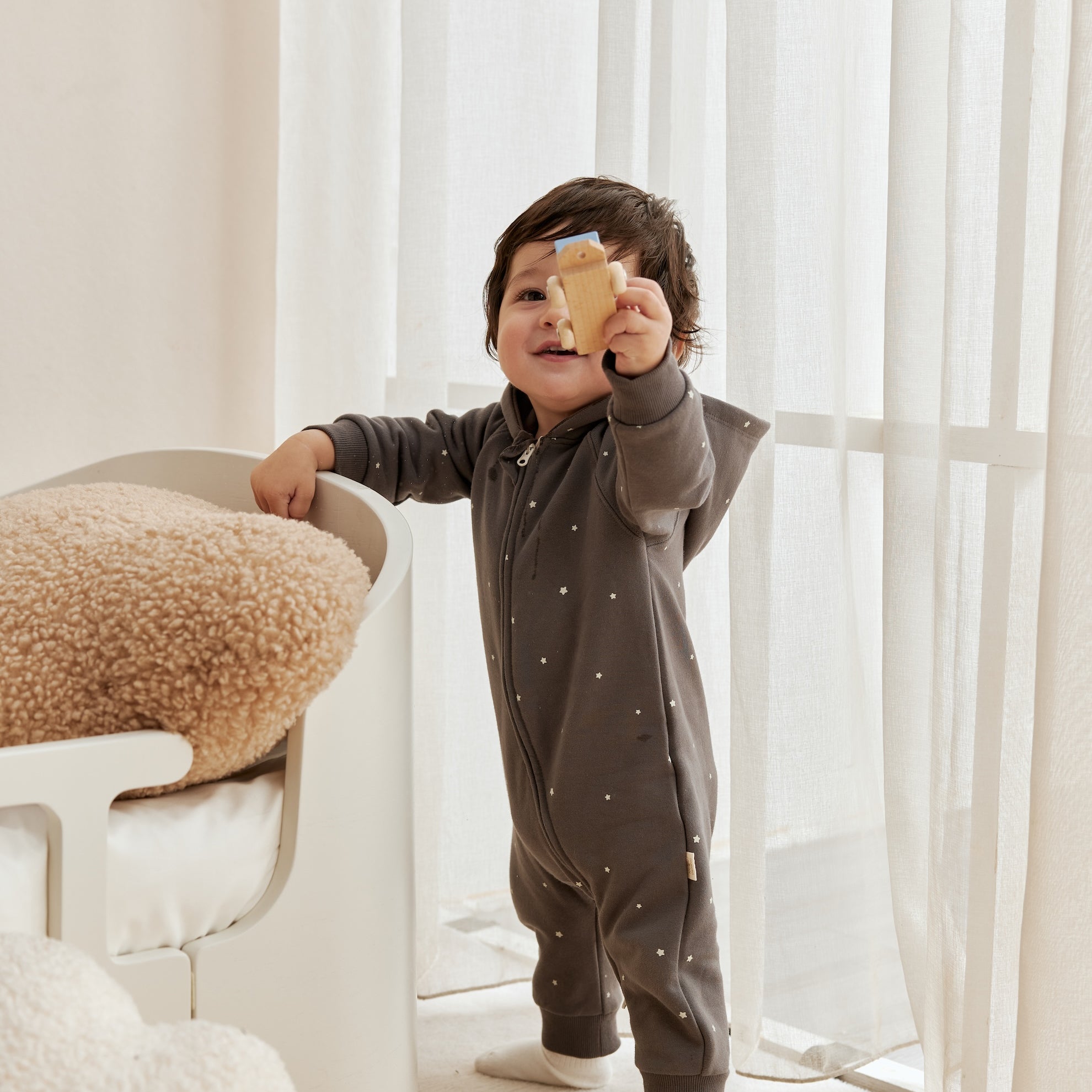 Child in a dark gray onesie holding a yellow object in a bright room with white curtains.
