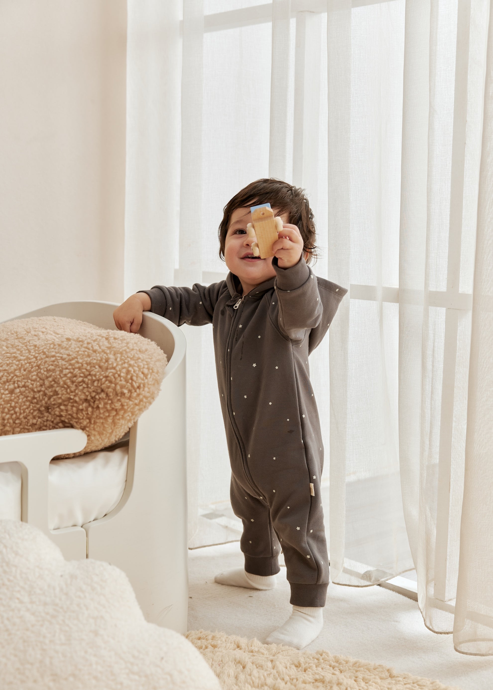 Child in a dark gray onesie holding a yellow object in a bright room with white curtains.