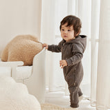 Child in a gray onesie standing in a bright room with white furniture and beige cushions.