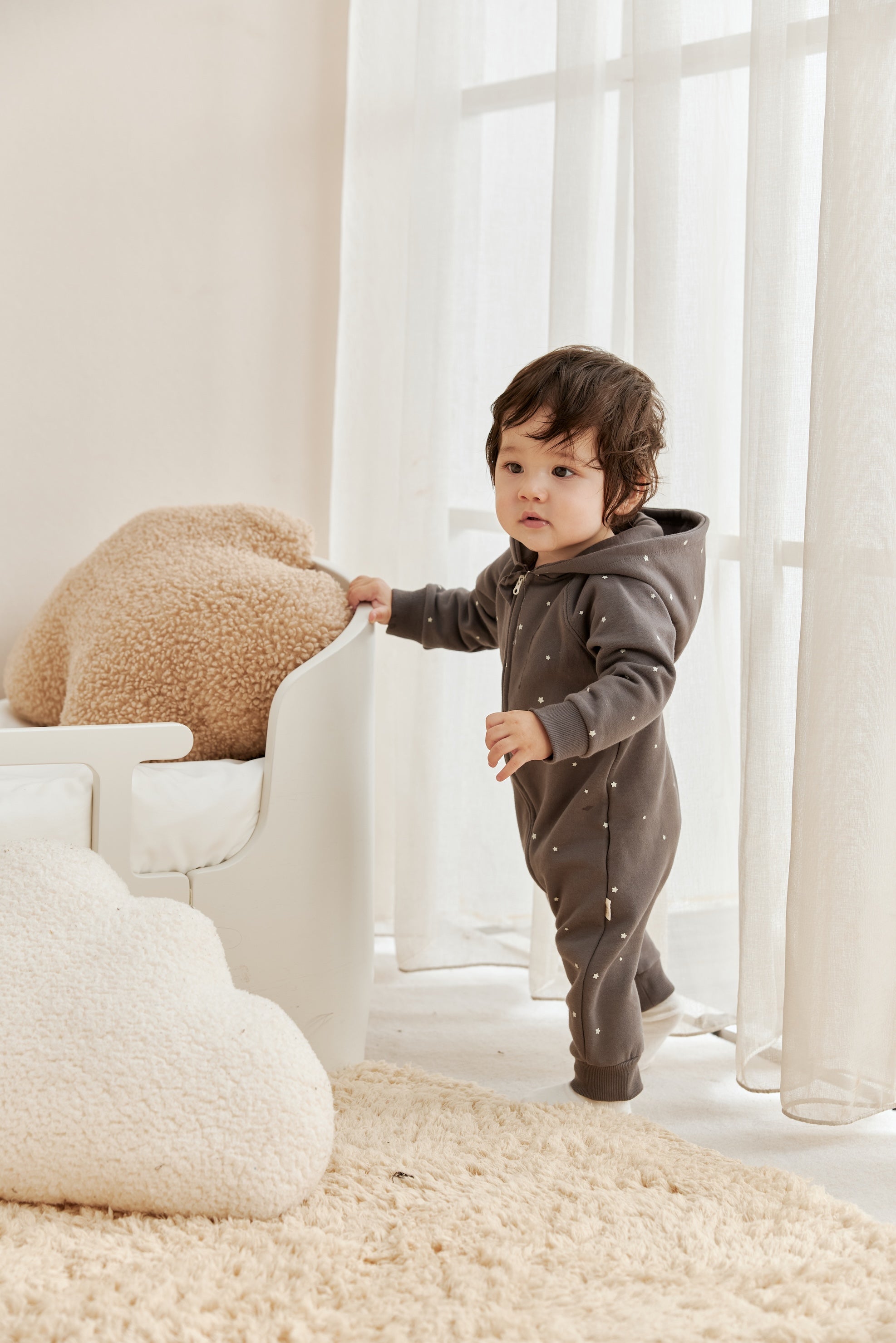 Child in a gray onesie standing in a bright room with white furniture and beige cushions.