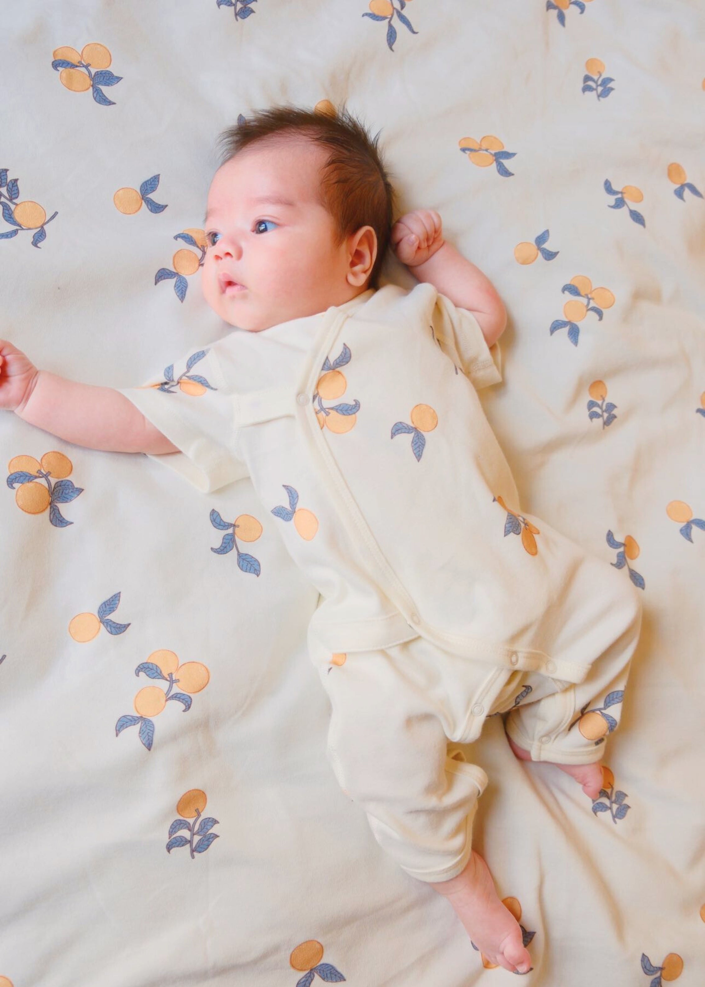 1. A cute baby resting on a bed, wearing a Clementine Organic Kimono Sleeper.
