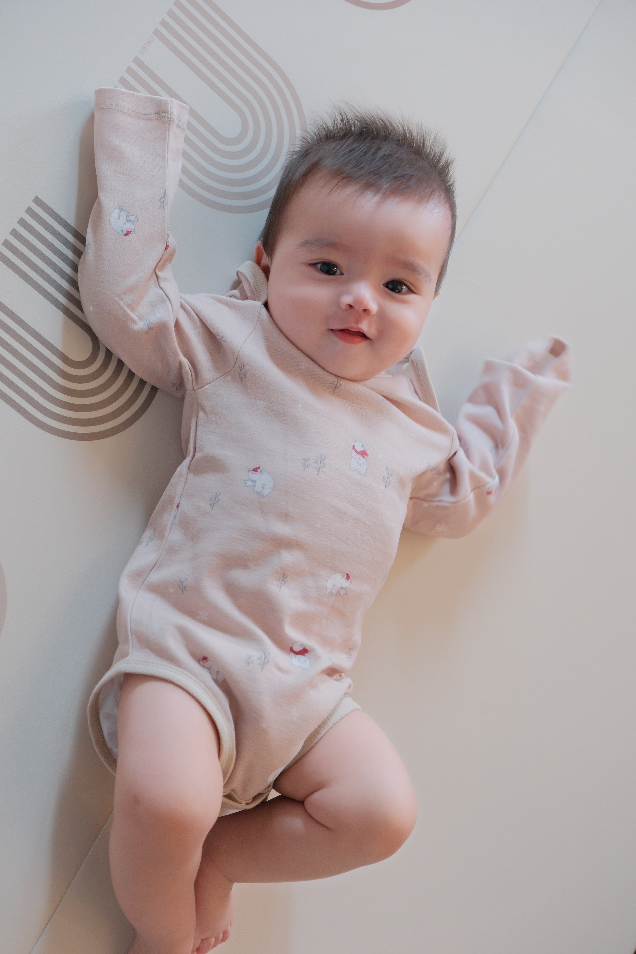 Baby in a light pink onesie lying on a white floor with a patterned rug.