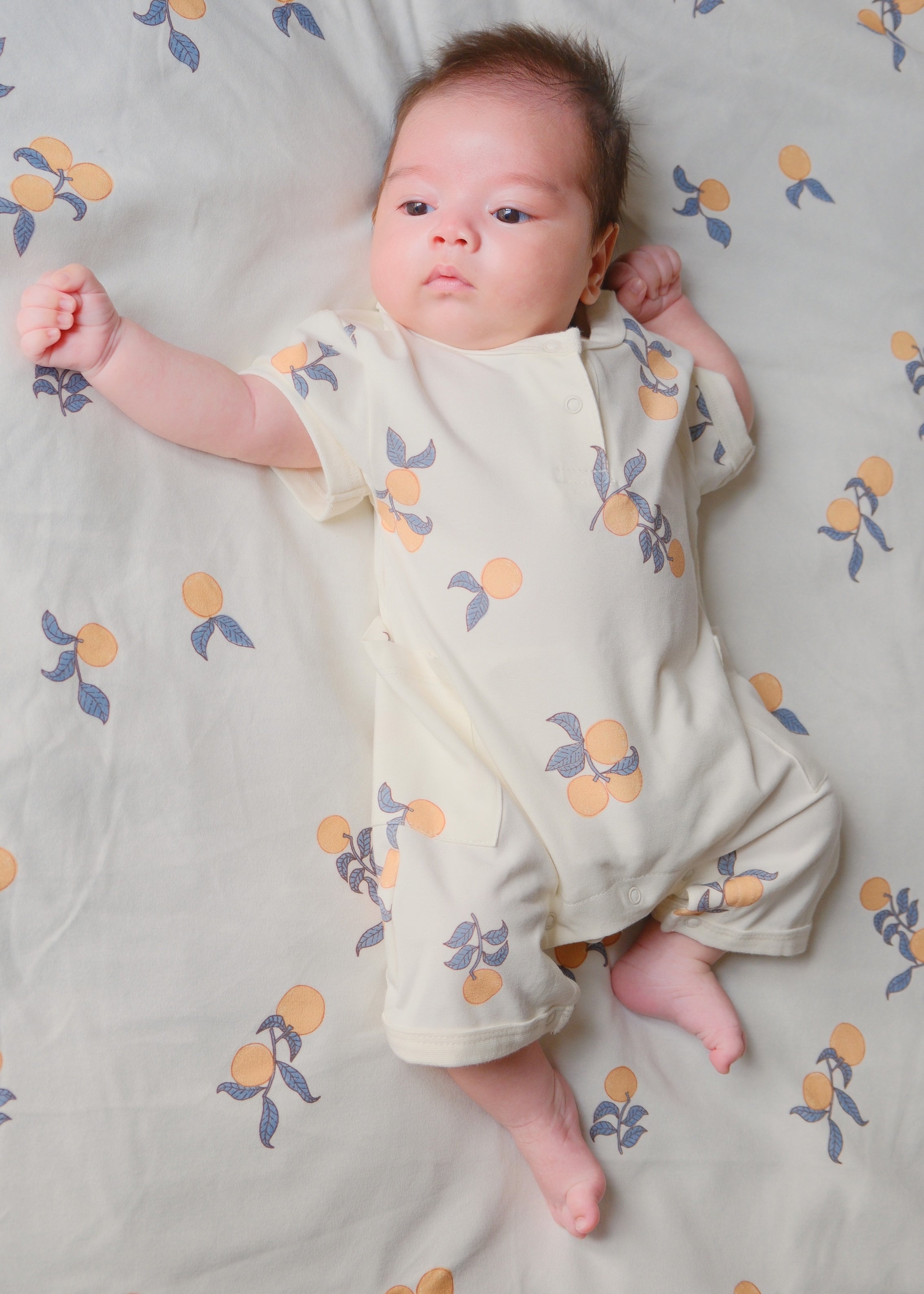 A baby in a orange print bed, wearing a cute collar tank romper in clementine color, looking cozy and adorable.
