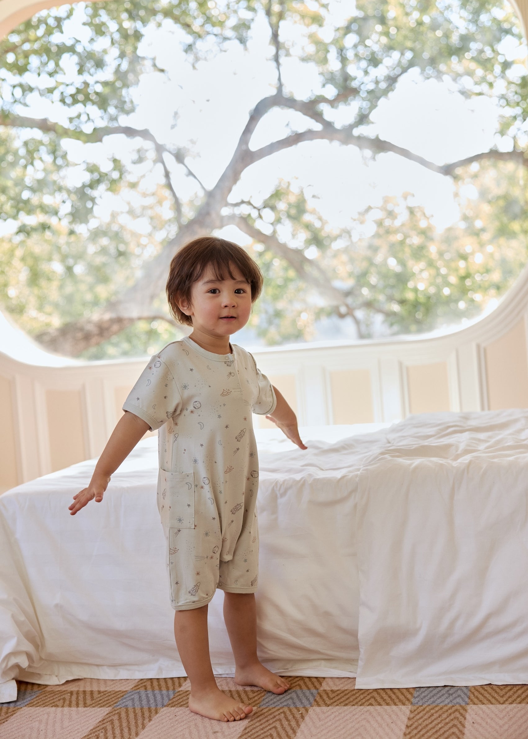 A young child sitting on a bed in a bright room, wearing a space print romper, with large windows and trees outside