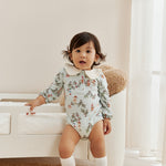Child wearing a floral onesie standing in a room with a white couch and light-colored walls.