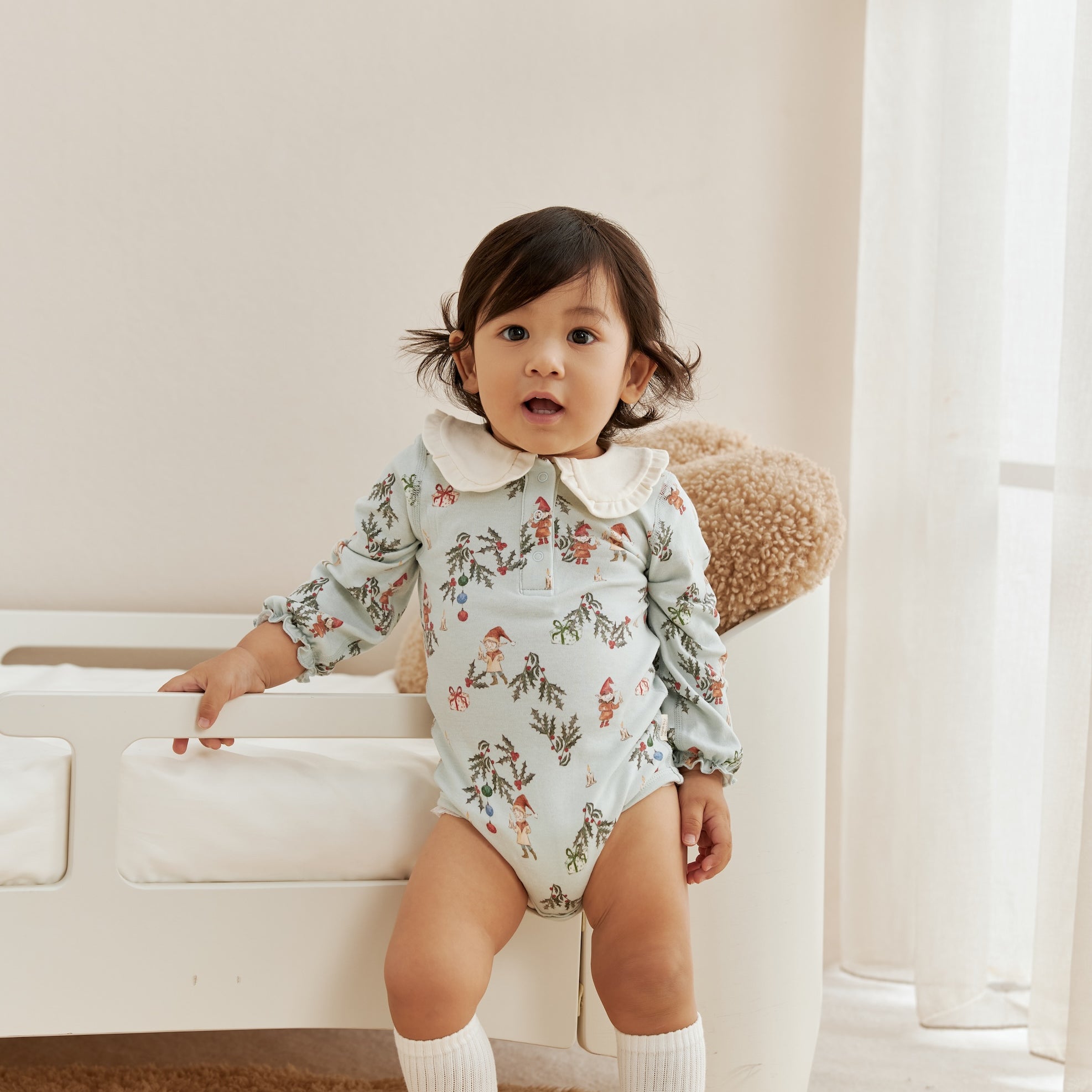 Child wearing a floral onesie standing in a room with a white couch and light-colored walls.