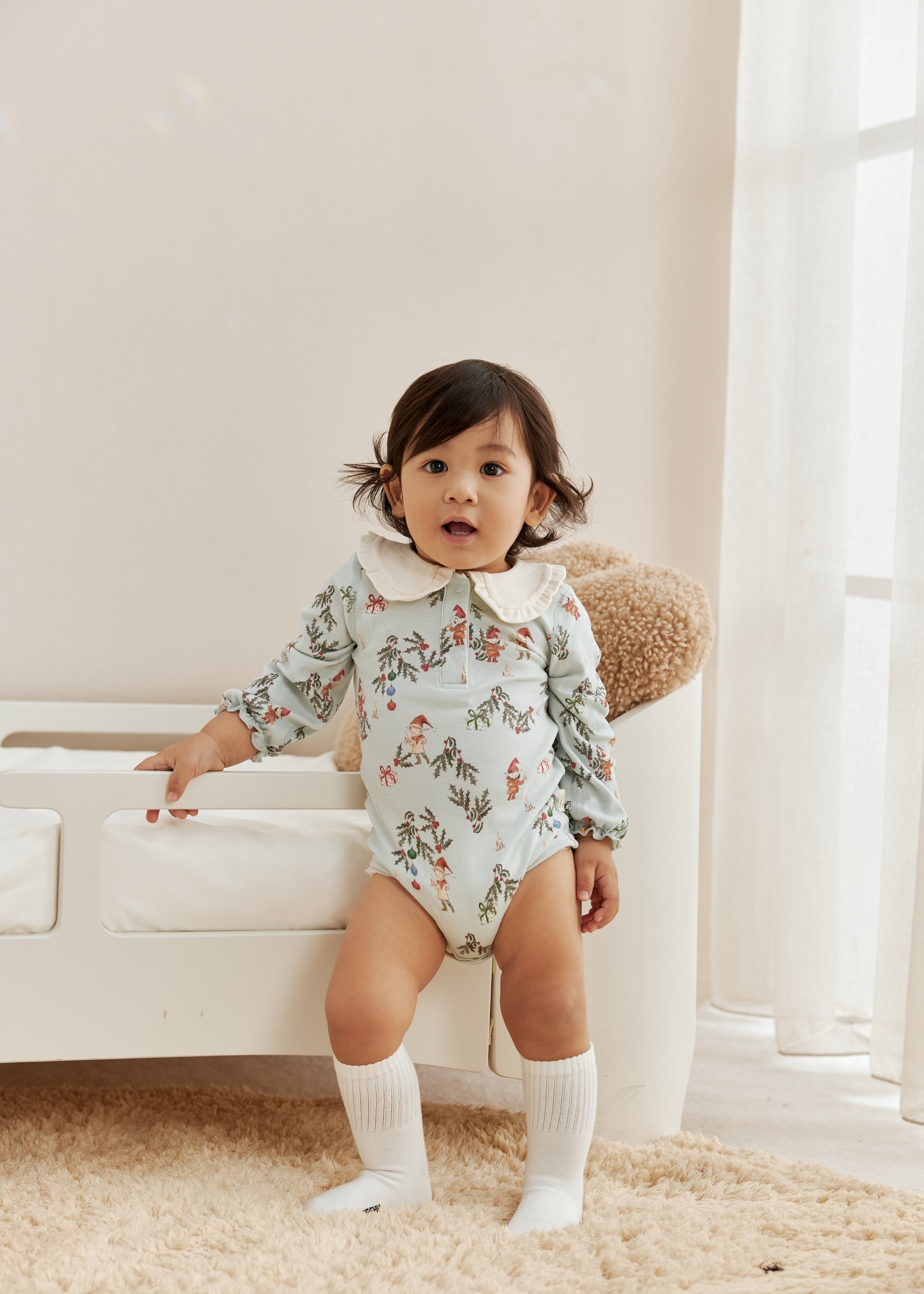 Child wearing a floral onesie standing in a room with a white couch and light-colored walls.