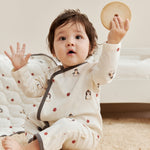 Baby in a white kimono sleeper with penguin design holding a wooden toy on a carpeted floor.