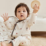 Baby in a white kimono sleeper with penguin design holding a wooden toy on a carpeted floor.